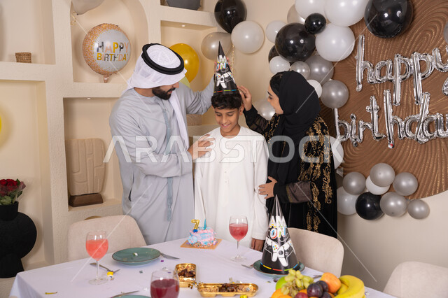 Sharing and giving gifts on occasions and birthdays, gestures of joy and happiness, feelings of love and tenderness in a happy family gathering, a family atmosphere filled with intimacy, balloons and a birthday cake decorated with candles, an Arab Gulf Emirati family having a good time at home.