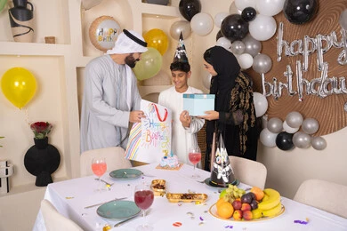 الاحتفال بأعياد الميلاد,  بلالين وكعكة يوم الميلاد المزينة بالشموع، التعبير عن المحبة والتقدير, أجواء أسرية مليئة بالألفة، عائلة عربية خليجية إماراتية تقضي أوقات ممتعه في المنزل، اب عربي خليجي اماراتي يقوم بمفجاءة و تقديم الهديه لابنه