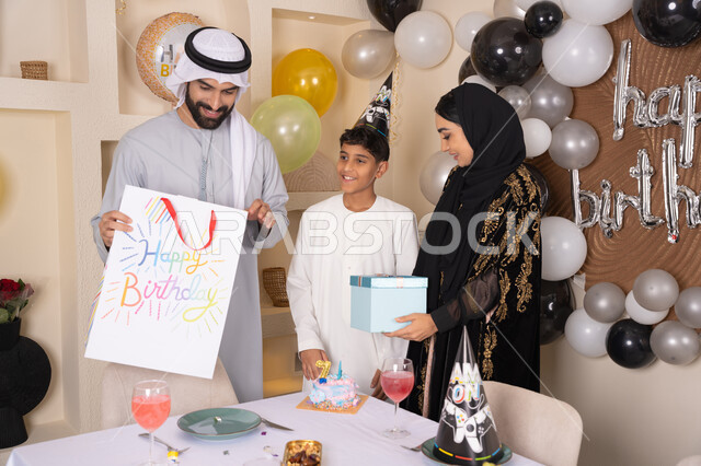 بلالين وكعكة يوم الميلاد المزينة بالشموع، الاحتفال بأعياد الميلاد, التعبير عن المحبة والتقدير, أجواء أسرية مليئة بالألفة، عائلة عربية خليجية إماراتية تقضي أوقات ممتعه في المنزل، اب عربي خليجي اماراتي يقوم بمفجاءة و تقديم الهديه لابنه