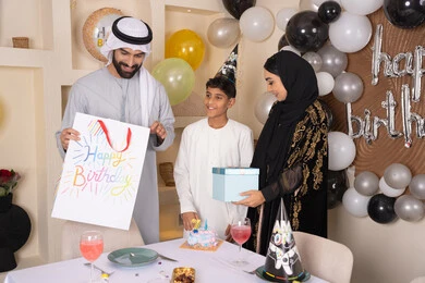 بلالين وكعكة يوم الميلاد المزينة بالشموع، الاحتفال بأعياد الميلاد, التعبير عن المحبة والتقدير, أجواء أسرية مليئة بالألفة، عائلة عربية خليجية إماراتية تقضي أوقات ممتعه في المنزل، اب عربي خليجي اماراتي يقوم بمفجاءة و تقديم الهديه لابنه