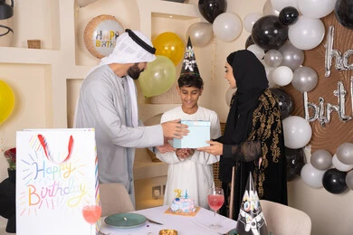 عائلة عربية خليجية إماراتية تقوم بالاحتفال وتقديم الهديه لابنهم ، أجواء أسرية مليئة بالألفة، الاحتفال بأعياد الميلاد,  بلالين وكعكة يوم الميلاد المزينة بالشموع، التعبير عن المحبة والتقدير
