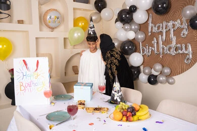 ام عربيه خليجيه اماراتية ترتدي العباءه والحجاب ، تقوم بالاحتفال و تقبيل ابنها بايماءات السعادة و السرور ، تعزيز روابط الامومه والحنان ، الاحتفال بأعياد الميلاد ضمن أجواء عائلية، تعليق الزينة والبالونات الملونة