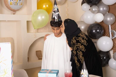 تعزيز روابط الامومه والحنان ، الاحتفال بأعياد الميلاد ضمن أجواء عائلية، تعليق الزينة والبالونات الملونة، ام عربيه خليجيه اماراتية ترتدي العباءه والحجاب ، تقوم بالاحتفال و تقبيل ابنها بايماءات السعادة و السرور