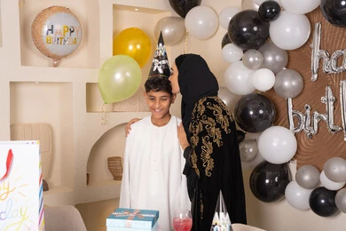 الاحتفال بأعياد الميلاد ضمن أجواء عائلية، تعليق الزينة والبالونات الملونة، ام عربيه خليجيه اماراتية ترتدي العباءه والحجاب ، تقوم بالاحتفال و تقبيل ابنها بايماءات السعادة و السرور ، تعزيز روابط الامومه والحنان
