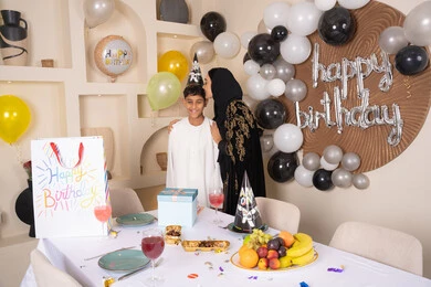 تعليق الزينة والبالونات الملونة، الاحتفال بأعياد الميلاد ضمن أجواء عائلية، ام عربيه خليجيه اماراتية ترتدي العباءه والحجاب ، تقوم بالاحتفال و تقبيل ابنها بايماءات السعادة و السرور ، تعزيز روابط الامومه والحنان