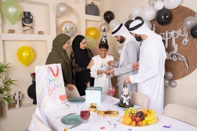 Balloons and a birthday cake decorated with candles, sharing and giving gifts on occasions and birthdays, gestures of joy and happiness, feelings of love and tenderness in a happy family gathering, an Arab Gulf Emirati family spending enjoyable times at home, a family atmosphere full of intimacy