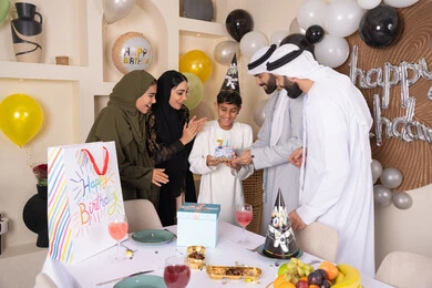 مشاركة وتقديم الهدايا بالمناسبات وأعياد الميلاد ، بلالين وكعكة يوم الميلاد المزينة بالشموع، إيماءات الفرح و السعادة، مشاعر الحب والحنان في اجتماع عائلي سعيد، عائلة عربية خليجية إماراتية تقضي أوقات ممتعه في المنزل، أجواء أسرية مليئة بالألفة