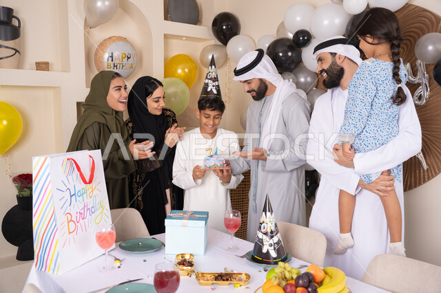 مشاركة وتقديم الهدايا بالمناسبات وأعياد الميلاد ، إيماءات الفرح و السعادة، بلالين وكعكة يوم الميلاد المزينة بالشموع، أجواء أسرية مليئة بالألفة،  مشاعر الحب والحنان في اجتماع عائلي سعيد,عائلة عربية خليجية إماراتية تقضي أوقات ممتعه في المنزل