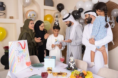 مشاركة وتقديم الهدايا بالمناسبات وأعياد الميلاد ، إيماءات الفرح و السعادة، بلالين وكعكة يوم الميلاد المزينة بالشموع، أجواء أسرية مليئة بالألفة،  مشاعر الحب والحنان في اجتماع عائلي سعيد,عائلة عربية خليجية إماراتية تقضي أوقات ممتعه في المنزل