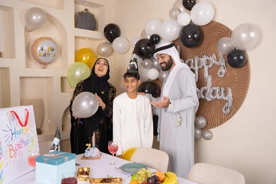 Balloons and a birthday cake decorated with candles, a family atmosphere full of intimacy, feelings of love and tenderness in a happy family gathering, an Arab Gulf Emirati family spending enjoyable times at home, gestures of joy and happiness, sharing and presenting gifts on occasions and birthdays