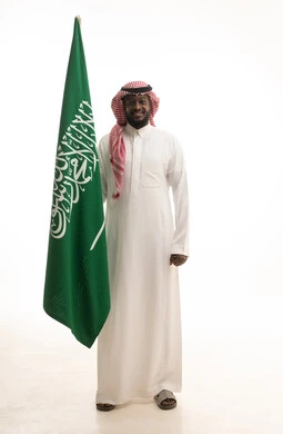 يوم العلم 11مارس، الاحتفال بذكرى اليوم الوطني السعودي 8 ربيع الأول, الفخر و الاعتزاز بالانتماء للمملكة, بورتريه لرجل عربي سعودي خليجي مبتسم يرتدي الشماغ والثوب التقليدي يحمل بيده علم السعودية , الجسم بالطول الكامل، خلفية بيضاء