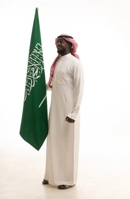 يوم العلم 11مارس، الاحتفال بذكرى اليوم الوطني السعودي 8 ربيع الأول, الفخر و الاعتزاز بالانتماء للمملكة, بورتريه لرجل عربي سعودي خليجي مبتسم يرتدي الشماغ والثوب التقليدي يحمل بيده علم السعودية , الجسم بالطول الكامل، خلفية بيضاء