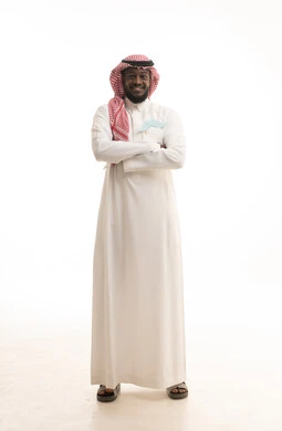 النظر الى الكاميرا و الوقوف بايدي متقاطعة ، تعزيز العافية البدنية للذكور، اليوم العالمي للرجل 19 نوفمبر، الإهتمام بمظهر الخارجي، الاحتفال بالمناسبات العالمية، بورتريه لرجل عربي خليجي سعودي يرتدي الثوب التقليدي والشماغ يحمل عود خشبي عليه قصاصات ورقية على شكل شارب، الطول الكامل للجسم، خلفية بيضاء