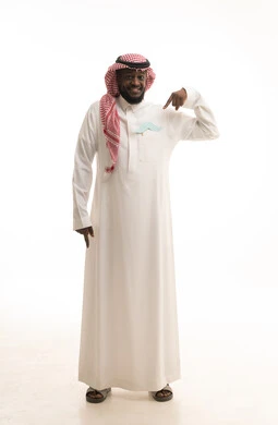 العناية بالصحة البدنية للرجال، مفهوم القوة والرجولة، إيماءات الثقة والاعتزاز بالنفس، بورتريه لرجل عربي خليجي سعودي يرتدي الثوب التقليدي والشماغ يضع في جيبه قصاصة ورقية على شكل شارب ويقوم بالإشارة اليها، صورة لكامل الجسم، خلفية بيضاء