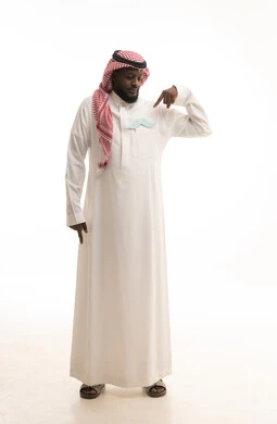 مفهوم القوة والرجولة، إيماءات الثقة والاعتزاز بالنفس، الاهتمام بالصحة الجسدية للذكور، بورتريه لرجل عربي خليجي سعودي يرتدي الثوب التقليدي والشماغ يضع في جيبه قصاصة ورقية على شكل شارب ويقوم بالإشارة اليها، صورة لكامل الجسم، خلفية بيضاء