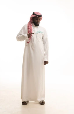 Attention to the physical health of males, gestures of confidence and self-esteem, the concept of strength and masculinity, a portrait of an Arab Gulf Saudi man wearing traditional attire and a ghutrah, placing a paper cutout in the shape of a mustache in his pocket and pointing to it, full-body image, white background.