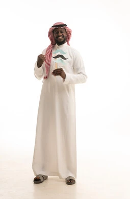 Gestures of victory and triumph, celebrating global occasions, focusing on men's health, maintaining fitness and muscle strength, International Men's Day on November 19, a portrait of an Arab Gulf Saudi man wearing traditional attire and a ghutrah holding a paper cutout in the shape of a mustache, full body length, white background.