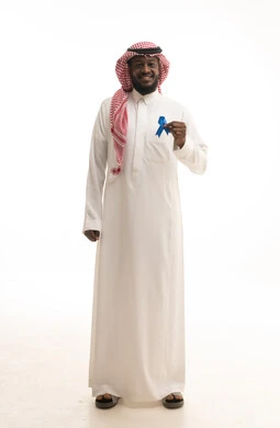 The importance of early detection of diseases, following medical guidelines, and not neglecting physical health, a portrait of a Saudi Arabian Gulf man wearing traditional attire and a shemagh holding a blue ribbon for prostate cancer awareness, full-body, white background.