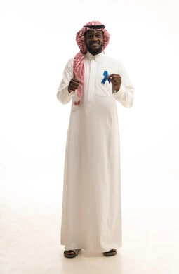 الكشف المبكر عن سرطان البروستات، حملات التوعية والتنبيه بعدم إهمال الصحة الجسدية، رفع اليد بإيماءات الاعجاب والرضا، بورتريه لشاب عربي خليجي سعودي يرتدي الزي التقليدي يمسك بيدية شريط أزرق للتوعية بالأمراض، صورة لكامل الجسم، خلفية بيضاء