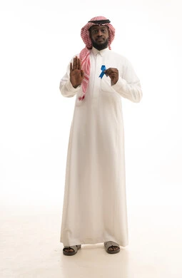 الوقوف باستقامة والنظر إلى الكاميرا ورفع اليد بإيماءات التحذير والتنبيه، حملات التوعية والإرشاد بعدم إهمال الصحة الجسدية، بورتريه لرجل عربي خليجي سعودي يرتدي الزي التقليدي يمسك بيدية شعار الكشف المبكر عن سرطان البروستات، صورة لكامل الجسم، خلفية بيضاء