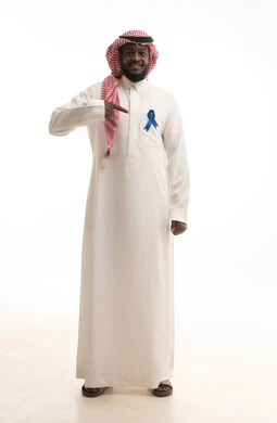 حملات إرشادية للانتباه للصحة، أهمية الكشف المبكر عن السرطانات،، بورتريه لرجل عربي خليجي سعودي يرتدي الثوب التقليدي والشماغ يقوم بالإشارة الى الشريط الأزرق للدلالة على سرطان البروستات، الجسم بالطول الكامل، خلفية بيضاء