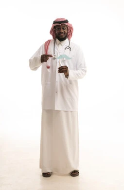 The reference to the paper with gestures of joy and pride, the annual International Men's Day, the concept of medicine and healthcare, a portrait of an Arab Gulf Saudi doctor wearing a ghutrah and a medical coat with a stethoscope, holding a wooden stick in one hand with a paper cutout shaped like a mustache, full body length, white background.