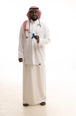 القيام بإجراءات الوقاية من السرطانات، تقديم الخدمات الصحية للمرضى، حملات التوعية والتنبيه، بورتريه لطبيب عربي خليجي سعودي يرتدي الشماغ و المعطف الطبي والسماعات الطبية يحمل بيده شعار الكشف المبكر عن سرطان البروستات، الطول الكامل للجسم، خلفية بيضاء