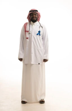 القيام بإجراءات الوقاية من السرطانات، تقديم الخدمات الصحية للمرضى، حملات التوعية والتنبيه، بورتريه لطبيب عربي خليجي سعودي يرتدي الشماغ و المعطف الطبي والسماعات الطبية يقف باستقامة يحمل بيده شعار الكشف المبكر عن سرطان البروستات، الطول الكامل للجسم، خلفية بيضاء