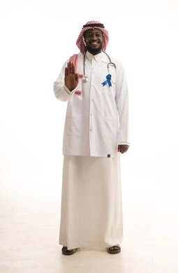 Standing upright, looking at the camera, and raising the hand with warning and alert gestures, awareness and guidance campaigns not to neglect physical health, a portrait of an Arab Gulf Saudi doctor wearing a ghutrah and a lab coat with the logo for early detection of prostate cancer on his chest, full-body image, white background.