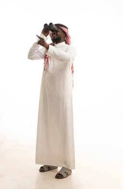 إيماءات الاستمتاع و السرور, حب الاستكشاف و المغامرة, بورتريه لرجل عربي خليجي سعودي يرتدي الثوب التقليدي و الشماغ يحمل بيده منظار ثنائي العينين يشير بإصبع السبابة للأعلى ينظر إلى شيئ ما, رؤية الاشياء البعيدة عبر التلسكوب, صورة الجسم بالطول الكامل, خلفية بيضاء