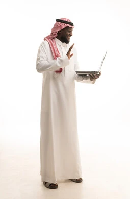 رفع كف اليد بتعابير السلام و التحية، إجراء مكالمة فيديو بإستخدام جهاز الحاسوب المحمول، بورتريه لرجل عربي خليجي سعودي يرتدي الزي التقليدي و الشماغ يتصفح الإنترنت، إستخدام اجهزة تقنية حديثة ، العمل عن بعد ، الجسم بالطول الكامل، خلفية بيضاء