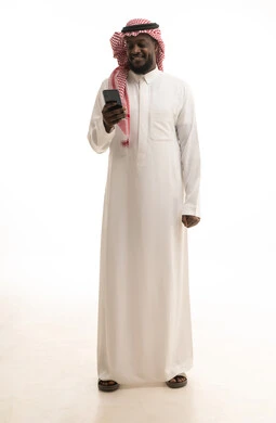 العمل عن بعد وانجاز المهام المطلوبة، استخدام جهاز تقني حديث ومتطور، تصفح مواقع التواصل الاجتماعي، بورتريه لرجل عربي خليجي سعودي يرتدي الشماغ والثوب التقليدي يمسك بيده الهاتف المحمول، الدردشة والتواصل مع الأهل و الأصدقاء، الجسم بالطول الكامل، خلفية بيضاء