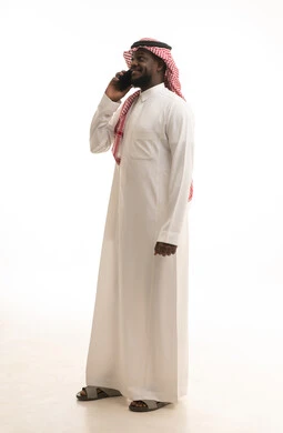 Making a phone call via mobile, communicating and talking with family and friends, a portrait of a Saudi Arabian Gulf man wearing traditional attire and a ghutrah holding a mobile phone in his hand, a full-body image, gray background.