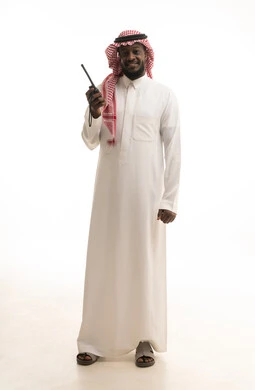 المتابعة والإشراف على خطط العمل، الإستجابة لحالات الطوارئ، بورتريه لرجل عربي خليجي سعودي يرتدي الثوب التقليدي والشماغ يقوم باستخدم جهاز الارسال والاستقبال اللاسلكي، مفهوم ادارة المشاريع عن بعد، صورة لكامل الجسم، خلفية بيضاء
