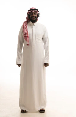 التواصل مع الزبائن لحل المشكلات والشكاوي، استخدام سماعات الرأس والتقنيات المتطورة في العمل، الرد على الاستفسارات والاستعلامات, بورتريه لرجل عربي خليجي سعودي يرتدي الشماغ والثوب التقليدي يضع الهيدفون يعمل في قسم خدمة العملاء، صورة الجسم بالكامل، خلفية بيضاء