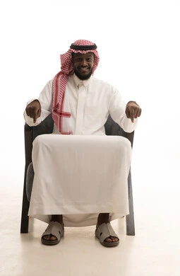 Expressions of specification and emphasis, signals that define this place (here), pointing to something, a portrait of a Saudi Arabian Gulf man wearing traditional attire and a ghutrah sitting on a chair and pointing down with his hands, white background.