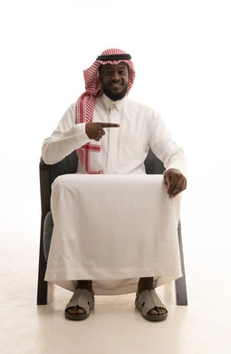 Indicators that define this place (here), expressions of specification and affirmation, pointing to something, a portrait of a Gulf Arab Saudi man wearing traditional attire and a ghutrah sitting on a chair and pointing with his index finger to the right, white background.