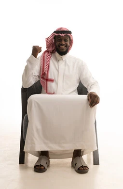 الاندماج بتشجيع كرة القدم, إيماءات الفوز والانتصار, بورتريه لرجل عربي خليجي سعودي مبتسم يرتدي الشماغ والثوب التقليدي يجلس على كرسي يرفع يديه بتفاعل وحماس, خلفية بيضاء