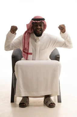 إيماءات الفوز والانتصار, بورتريه لرجل عربي خليجي سعودي مبتسم يرتدي الشماغ والثوب التقليدي يجلس على كرسي يرفع يديه بتفاعل وحماس, الاندماج بتشجيع كرة القدم, خلفية بيضاء