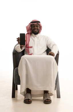 A blank black screen displayed on a mobile phone, looking at the camera with gestures of joy and happiness, a portrait of a smiling Arab Saudi Gulf man wearing traditional attire and a shemagh sitting on a comfortable chair holding a mobile phone, using advanced modern technology, white background.