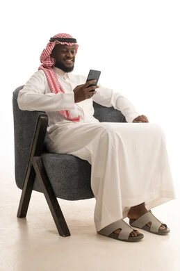 Browsing social media on a mobile phone, completing work tasks remotely, enjoying and entertaining oneself using modern technology, gestures of happiness and joy, a portrait of an Arab Gulf Saudi man wearing traditional attire and a ghutrah sitting on a comfortable chair using a mobile phone, white background.