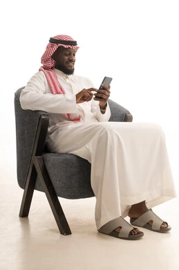 المتعة والتسلية باستخدام التقنية الحديثة، تصفح مواقع التواصل الاجتماعي عبر الجوال، إنجاز مهام العمل عن بعد، بورتريه لرجل عربي خليجي سعودي يرتدي الثوب التقليدي و الشماغ يجلس على كرسي مريح يقوم باستخدام الهاتف المحمول، خلفية بيضاء