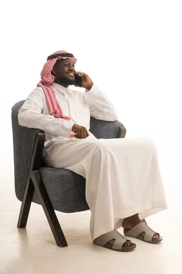 The feeling of happiness upon hearing some news, making phone calls via mobile phone, communicating with family and friends, a portrait of a smiling Arab Saudi Gulf man wearing traditional attire and a shemagh sitting on a comfortable chair using a mobile phone, using modern technological means in professional life, white background.