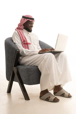 The use of modern technological devices, a portrait of a Saudi Arabian Gulf man wearing a thobe and ghutrah sitting on a chair with a laptop in front of him, chatting with family and friends, browsing social media platforms, computer applications, white background.