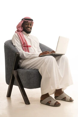 استخدام الأجهزة التقنية الحديثة, بورتريه لرجل عربي خليجي سعودي يرتدي الثوب والشماغ يجلس على كرسي ويضع امامه حاسب محمول، الدردشة مع الأهل والأصدقاء, تصفح برامج التواصل الاجتماعي، تطبيقات الأجهزة الحاسوبية، خلفية بيضاء
