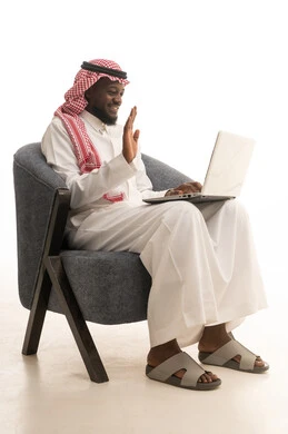 Making a video call over the internet, raising the hand with expressions of happiness and joy, a portrait of a Gulf Arab Saudi man wearing a thobe and shemagh sitting on a chair with a laptop in front of him, chatting with family and friends, browsing social media, computer applications, white background.