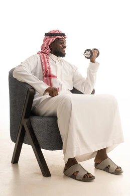 رفع الأوزان الحديدية الثقيلة، ممارسة التمارين الرياضية لتقوية وبناء العضلات، نشاط بدني للمحافظة على الصحة واللياقة الجسدية، اتباع أسلوب حياة صحي، بورتريه لرجل عربي خليجي سعودي يرتدي الزي التقليدي و الشماغ  يجلس على كرسي يحمل الدمبلز، خلفية بيضاء