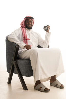 نشاط بدني للمحافظة على الصحة واللياقة الجسدية، ممارسة التمارين الرياضية لتقوية وبناء العضلات، اتباع أسلوب حياة صحي، بورتريه لرجل عربي خليجي سعودي يرتدي الزي التقليدي و الشماغ  يجلس على كرسي يحمل الدمبلز، رفع الأوزان الحديدية الثقيلة، خلفية بيضاء