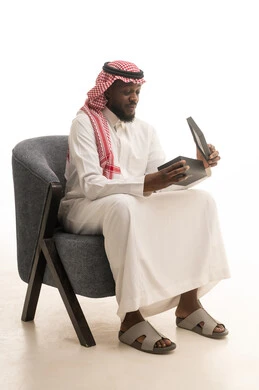 The concept of gratitude and appreciation, giving gifts during holidays and special occasions, expressing love, respect, and admiration, a portrait of a Saudi Arabian Gulf man wearing traditional attire and a ghutrah sitting on a comfortable chair opening a gift box with gestures of joy and happiness, white background.