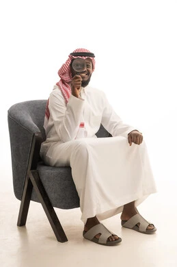 مفهوم الفحص والاستكشاف، أدوات بصرية للتدقيق والتركيز، توضيح الصورة والرؤية عن قرب، بورتريه لرجل عربي خليجي سعودي يرتدي الثوب التقليدي و الشماغ يجلس على كرسي مريح يحمل بيده عدسة مكبرة ينظر من خلالها بإيماءات التركيز والتمعن، خلفية بيضاء